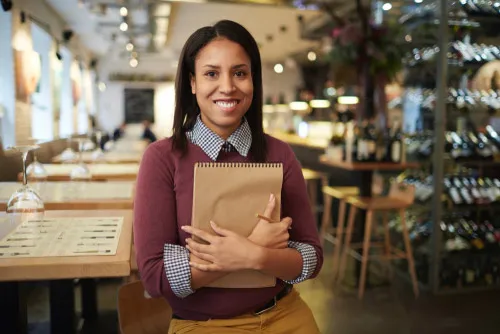 A restaurant manager stands in front of an empty restaurant interior while holding a large notepad